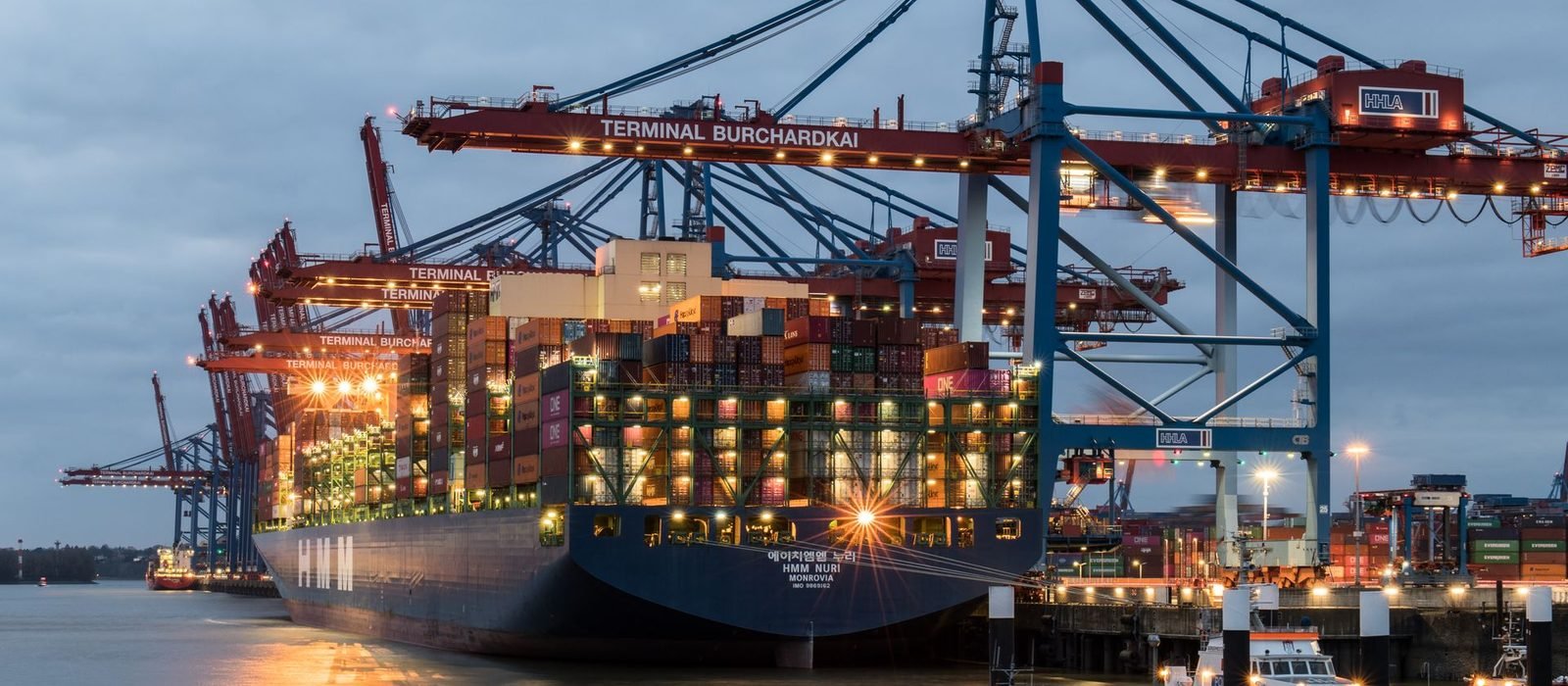 A container ship berthed at a terminal at dusk, gantry cranes lit above the stacked containers